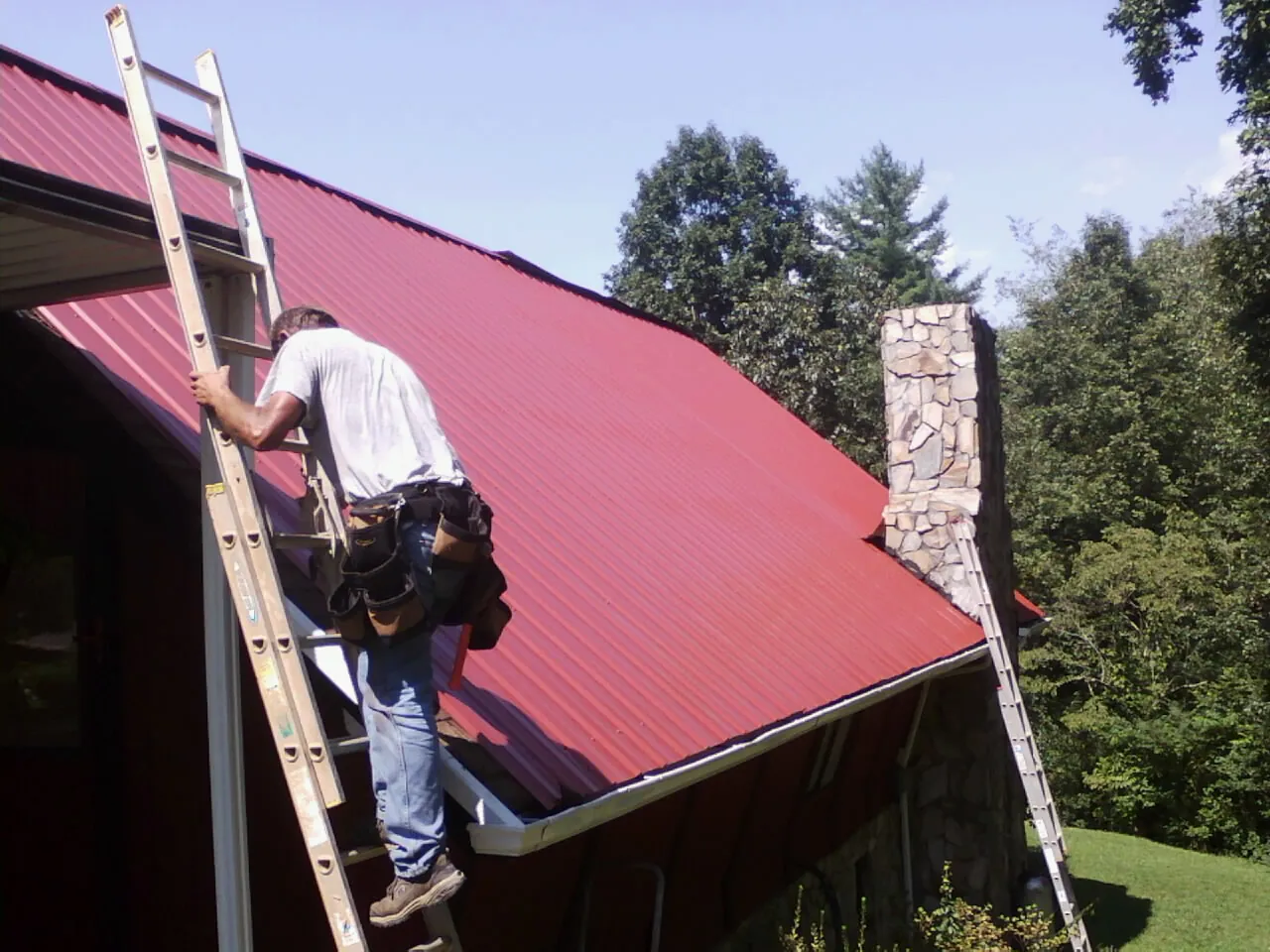Roofer on a ladder beside a red metal roof and stone chimney — Soffit & Fascia Repair by Roofing Brothers in Rougemont